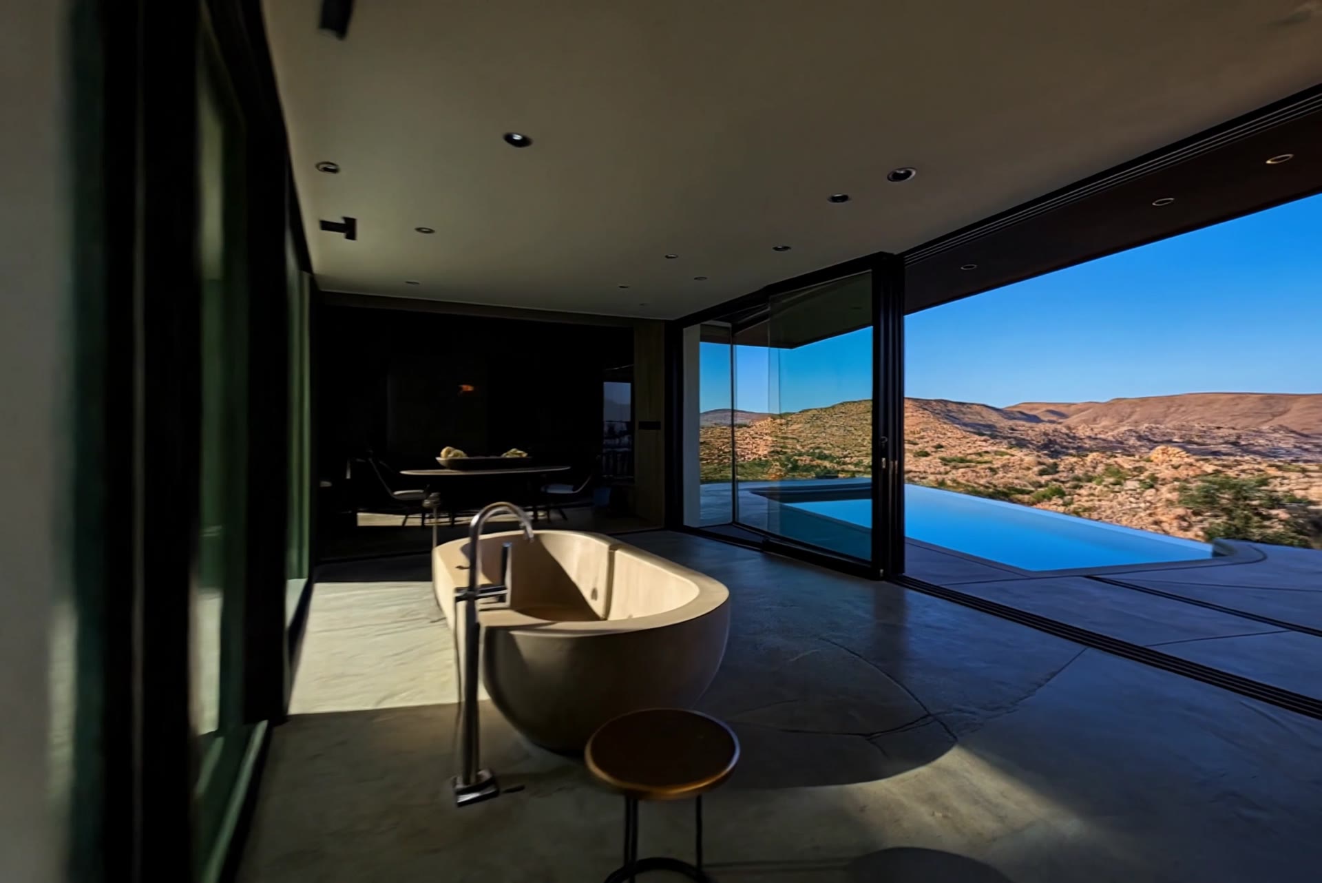Open-plan desert retreat with a freestanding stone bathtub and floor-to-ceiling glass opening onto an infinity pool and canyon views